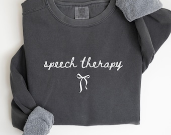 May include: A dark gray sweatshirt with the words "speech therapy" in white script and a white bow graphic. The sweatshirt has a fleece-lined collar and cuffs. The word "PEPPER" is printed in white at the bottom.