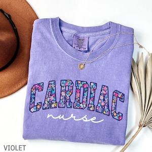 May include: A violet Comfort Colors t-shirt with the words "CARDIAC nurse" in a floral design. A gold necklace with a pendant is draped across the shirt. A brown hat is visible in the upper left corner.