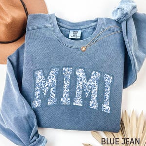 May include: A blue jean-colored sweatshirt with the word "MIMI" in large, white, floral-patterned letters across the front. A gold necklace with a pendant rests on the sweatshirt. The text "BLUE JEAN" is at the bottom.