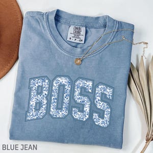 Peut inclure: Un t-shirt bleu jean avec le mot "BOSS" en grandes lettres à motifs floraux. Un collier en or avec un pendentif repose sur le t-shirt. Le t-shirt a un col rond et des manches courtes. L'étiquette indique "Comfort Colors".