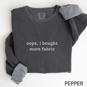 May include: A dark gray sweatshirt with the text "oops, i bought more fabric" in white. The sweatshirt has a crew neck and long sleeves with gray, fuzzy cuffs. The word "PEPPER" is printed in black at the bottom.