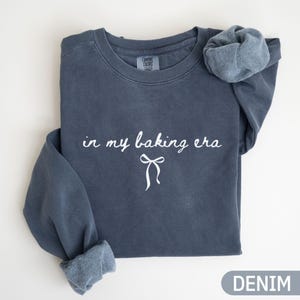 Könnte beinhalten: Ein denimblaues Sweatshirt mit dem weißen Schriftzug "in my baking era" und einer weißen Schleifengrafik. Das Sweatshirt hat hochgekrempelte Ärmel und ein "Comfort Colors"-Etikett.