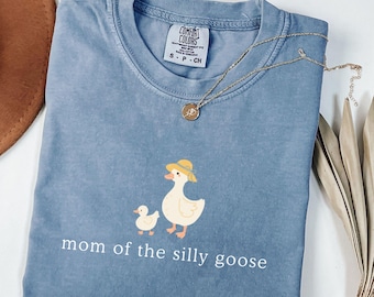 May include: A blue jean Comfort Colors t-shirt with a graphic of a mother goose and gosling wearing hats. The text "mom of the silly goose" is printed below the graphic. A gold necklace is draped across the shirt.