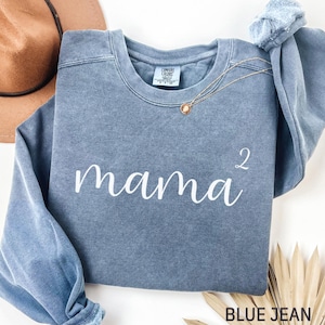 May include: Blue jean-colored sweatshirt with the word "mama²" in white script. A gold necklace with a pendant rests on the shirt. The sweatshirt is folded, with a brown hat visible in the background.