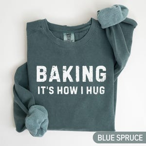 May include: Blue spruce-coloured sweatshirt with the text "BAKING IT'S HOW I HUG" in white block letters. The sweatshirt is a crew neck style with long sleeves. The words "BLUE SPRUCE" are at the bottom right.