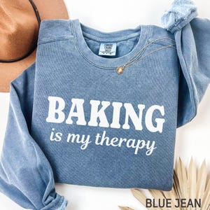 May include: A blue sweatshirt with the words "BAKING is my therapy" printed in white. A gold necklace with a pendant is on the shirt. A brown hat is in the background.