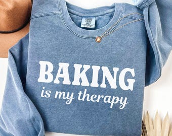 May include: Blue jean-colored sweatshirt with the text "BAKING is my therapy" in white. A gold necklace with a pendant rests on the shirt. A brown hat is visible in the background.