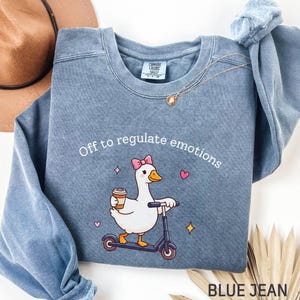 May include: A blue jean coloured sweatshirt with the slogan "Off to regulate emotions." The graphic shows a white goose with a pink bow on a scooter, holding a coffee cup. The sweatshirt has a crew neck and long sleeves.