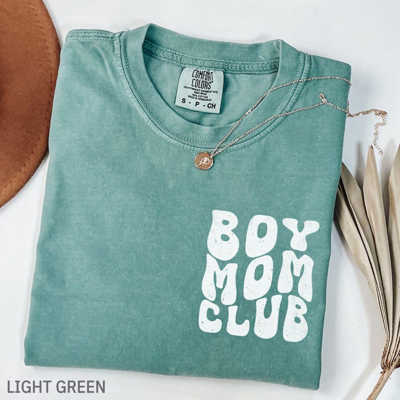 以下が含まれることがあります： 白文字で「BOY MOM CLUB」と書かれた、ライトグリーンのComfort Colors Tシャツ。ペンダント付きのシルバーネックレスがシャツの上にかけられています。シャツは折りたたまれており、背景には茶色の帽子が見えます。
