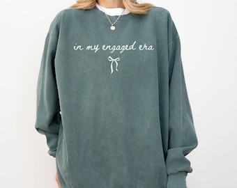 May include: Blue spruce-colored sweatshirt with the text "in my engaged era" and a bow graphic. The sweatshirt has long sleeves and a crew neck. The words "BLUE SPRUCE" are at the bottom of the image.