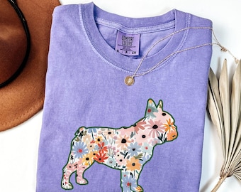 Puede incluir: Una camiseta de color violeta con un diseño floral de perro. La silueta del perro está llena de flores coloridas. Un collar dorado y un sombrero marrón son visibles en el fondo. La palabra "VIOLET" está impresa en la parte inferior.