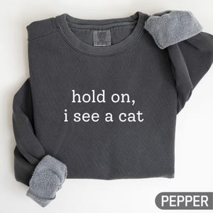 以下が含まれることがあります： 「hold on, i see a cat」の白文字が入った濃いグレーのスウェットシャツ。スウェットシャツは裏地がフリース素材で、「PEPPER」の文字が下部にグレーの長方形で表示されています。