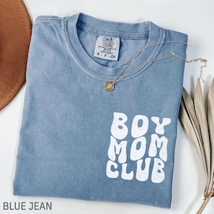 以下が含まれることがあります： 白い文字で「BOY MOM CLUB」と書かれた、青いジーンズカラーのTシャツ。ペンダント付きのゴールドネックレスがシャツの上にあります。シャツは白い面に折りたたまれています。