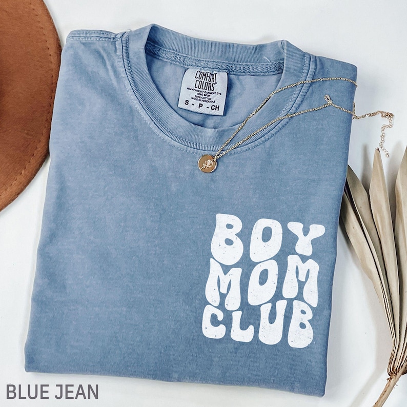 以下が含まれることがあります： 白い文字で「BOY MOM CLUB」と書かれた、青いジーンズカラーのTシャツ。ペンダント付きのゴールドネックレスがシャツの上にあります。シャツは白い面に折りたたまれています。