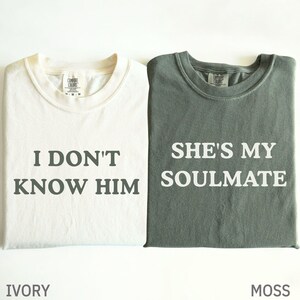 Puede incluir: Dos camisetas, una marfil y otra verde musgo, se muestran juntas. La camiseta marfil dice "I DON'T KNOW HIM" en texto verde oscuro. La camiseta verde musgo dice "SHE'S MY SOULMATE" en texto blanco. Ambas tienen cuello redondo.