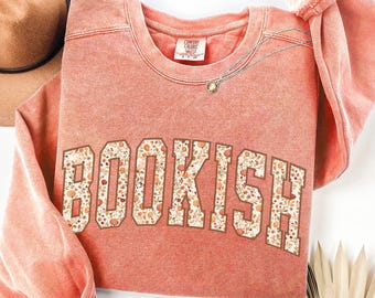 May include: Terracotta-colored sweatshirt with the word "BOOKISH" in a collegiate-style font. The letters are filled with a floral pattern of orange and cream-colored flowers and leaves. A gold necklace with a pendant is draped on the shirt.