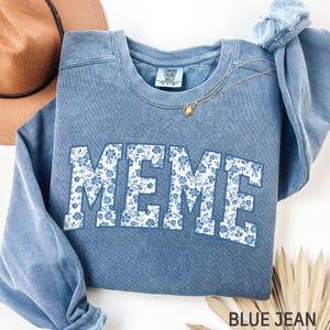 May include: A blue jean coloured sweatshirt with the word "MEME" in large, white, block letters. The letters are filled with a blue floral pattern. A gold necklace with a small pendant rests on the sweatshirt. The words "BLUE JEAN" are printed at the bottom.
