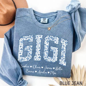 Könnte beinhalten: Ein blaues Jeans-Sweatshirt mit dem Namen "GIGI" in großen, weißen Buchstaben mit Blumenmuster. Darunter stehen die Namen Sophia, Oliver, James, Bella, Emma, Amelia und Max. Eine goldene Halskette und ein brauner Hut sind sichtbar.