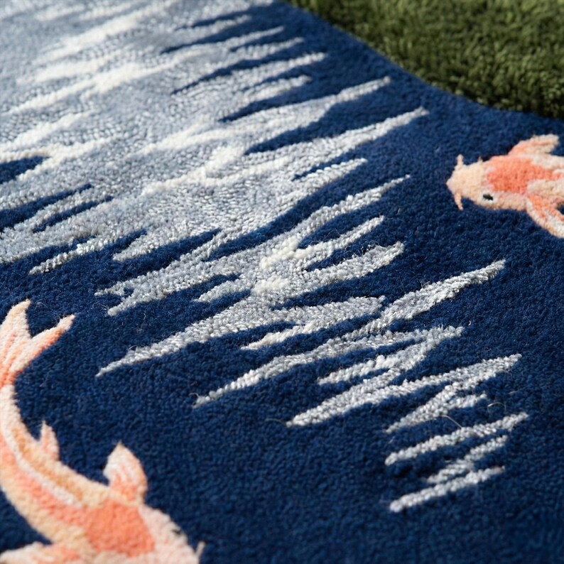 May include: Close-up of a blue rug with a design featuring white and gray wave-like patterns and two orange and white koi fish. The rug has a textured surface and a dark blue background.
