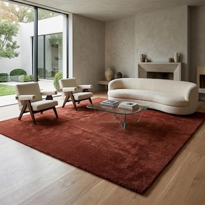 May include: A living room set featuring a large, curved, cream-colored sofa, two wooden armchairs with cream cushions, a glass-topped coffee table, and a large, textured, rust-colored area rug placed on light-colored wooden flooring.
