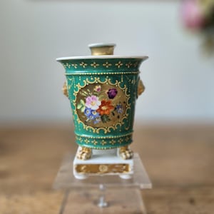 May include: A small, ornate green and gold porcelain vase with a lid. The vase features a floral design within a gold frame, with gold accents and small lion's head handles. It stands on a white base with gold feet.