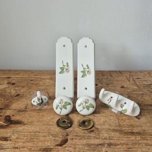 Op de afbeelding: Een verzameling witte keramische kastbeslagen met een bloemenrozenontwerp. De set bevat twee lange grepen, twee ronde knoppen, een kleine haak en een multi-haakrek. De hardware staat op een rustiek houten oppervlak.