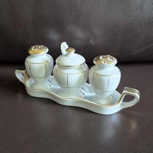 Op de afbeelding: Een witte porseleinen kruidenset met gouden details. De set bevat een zoutstrooier, een peperstrooier en een suikerpot met deksel, alles op een bijpassend dienblad met handvatten. Klassiek, elegant ontwerp.