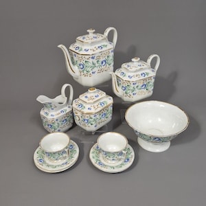 Puede incluir: Un juego de té de porcelana blanca con diseños florales azules y ribetes dorados. El juego incluye una tetera, una lechera, una azucarera, un cuenco y dos tazas de té con platillos. El juego de té está dispuesto sobre una superficie gris.