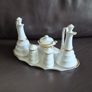 Op de afbeelding: Een witte porseleinen condimentenset met gouden rand. De set bevat twee olie- of azijnflessen, twee zoutvaatjes, een suikerpot met deksel en een serveerblad. Voor de eettafel.