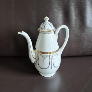 Op de afbeelding: Witte porseleinen theepot met een gebogen tuit, handvat en deksel. De theepot is versierd met gouden randen en blauwe en gouden accenten. De theepot staat op een bruin oppervlak.