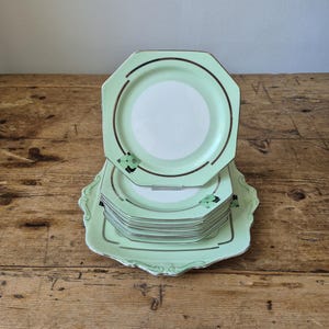 May include: A stack of octagonal light green plates with a white center and a silver metallic rim. The plates feature a small black and green geometric decal on the edge. The stack rests on a larger, matching light green octagonal serving tray.