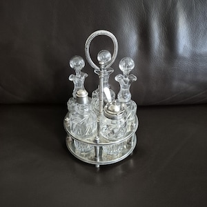 Może przedstawiać: Zestaw przypraw w stylu vintage, posrebrzany, z przezroczystymi szklanymi butelkami i słoiczkami. Zestaw zawiera centralny uchwyt, solniczkę i kilka butelek z ozdobnymi korkami. Zestaw jest ułożony na wielopoziomowej, okrągłej podstawie.