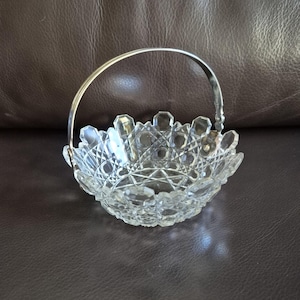 May include: A clear, cut-glass basket with a silver handle. The basket has a scalloped edge and a detailed, geometric pattern. The handle arches over the bowl, creating a decorative and functional design.