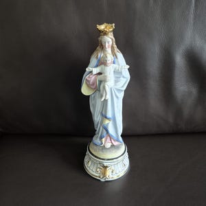 Peut inclure: Statuette en porcelaine de la Vierge Marie tenant l'enfant Jésus. La statue porte une robe bleu clair avec des détails dorés et une couronne dorée. Le socle est blanc avec des bordures dorées.
