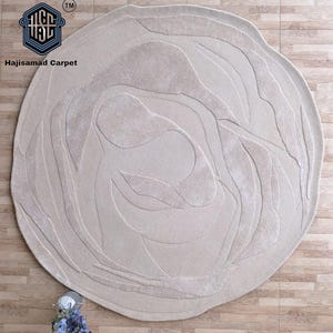 May include: A beige, round rug with a floral design. The rug features a raised pattern of curved lines and leaf-like shapes. The rug is placed on a wooden floor. The brand name "Hajisamad Carpet" is visible in the upper left corner.