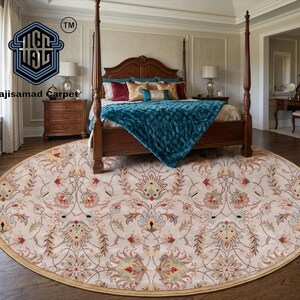 May include: A round beige rug with a floral pattern in red, blue, and gold tones. The rug is centered in a bedroom with a four-poster bed and wooden furniture. The text "Hajisamad Carpet" is visible.