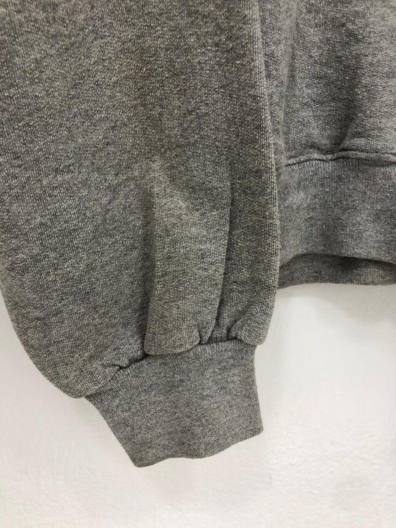 May include: Close-up of a gray sweatshirt sleeve. The cuff is ribbed and the fabric has a textured appearance. The sleeve is gathered at the cuff, creating a slight puff. The background is a plain white surface.
