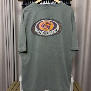 K&ouml;nnte beinhalten: Olivgr&uuml;nes T-Shirt mit einem gro&szlig;en, ovalen Aufdruck auf der Vorderseite. Die Grafik zeigt ein stilisiertes "G" in einem Kreis mit dem Wort "GRAMICCI" darunter. Das Shirt hat kurze &Auml;rmel und eine lockere Passform.