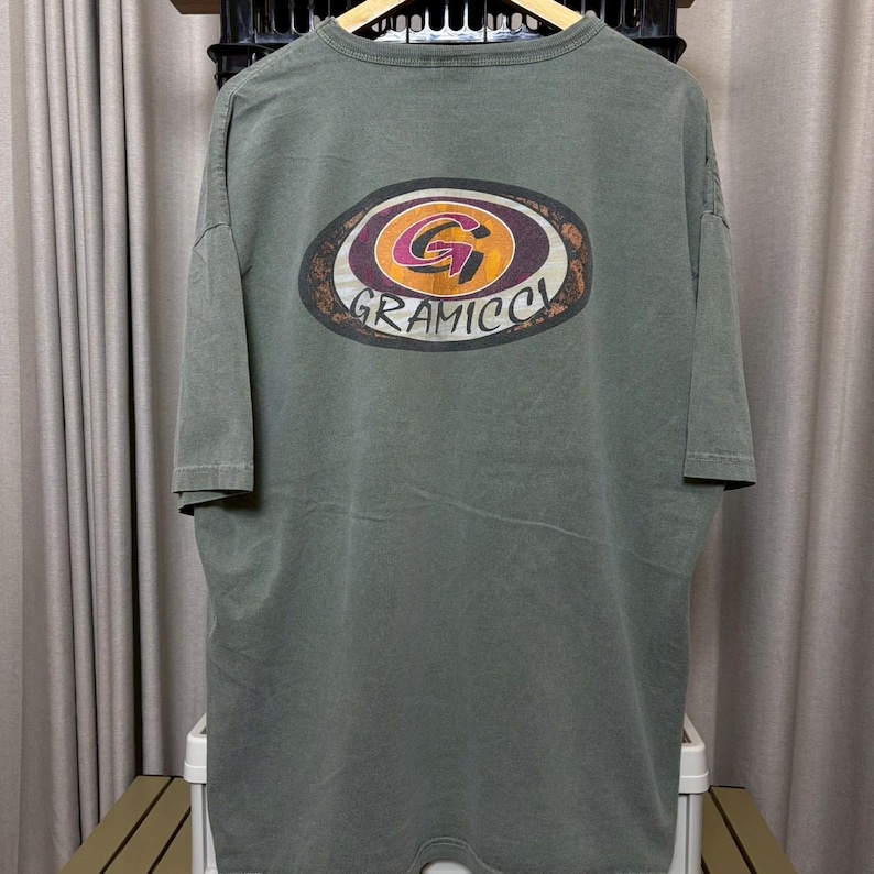 K&ouml;nnte beinhalten: Olivgr&uuml;nes T-Shirt mit einem gro&szlig;en, ovalen Aufdruck auf der Vorderseite. Die Grafik zeigt ein stilisiertes "G" in einem Kreis mit dem Wort "GRAMICCI" darunter. Das Shirt hat kurze &Auml;rmel und eine lockere Passform.