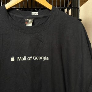 Könnte beinhalten: Ein schwarzes kurzärmeliges T-Shirt mit einem weißen Apple-Logo und dem Schriftzug "Mall of Georgia" auf der Brust. Das T-Shirt hat ein kleines weißes Etikett am inneren Halsausschnitt mit den handschriftlichen Maßen 59,7 mal 74,9 cm.