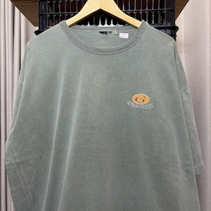 K&ouml;nnte beinhalten: Salbeigr&uuml;nes T-Shirt mit Rundhalsausschnitt und kurzen &Auml;rmeln. Das Shirt hat ein orange-gelbes Logo mit dem Wort "GRAMICCI" darunter. Das T-Shirt h&auml;ngt an einem Holzb&uuml;gel.