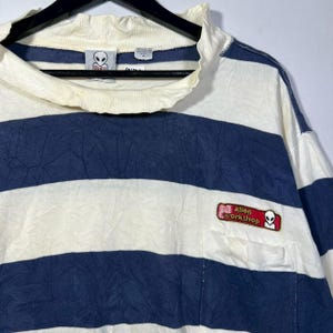 May include: Cream and navy blue striped long-sleeve shirt with a mock neck. The shirt features a small pocket with an "alien workshop" patch and an alien logo. The shirt is hanging on a black hanger.