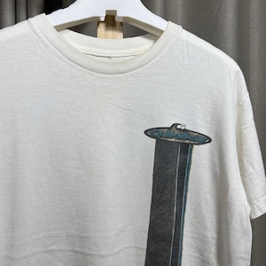 May include: White t-shirt with a crew neck and short sleeves. The shirt features a graphic of a stylized UFO with the text "Quiksilver" in a blue oval shape, above a dark gray vertical design.