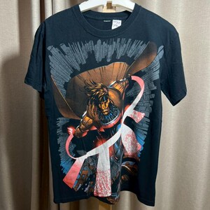 May include: Black t-shirt featuring a comic book character in action. The graphic shows a figure with a brown cape and a red and white design. The background has a cityscape design. The shirt is hanging on a white hanger.