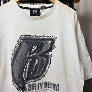 Könnte beinhalten: Ein cremefarbenes Kurzarm-T-Shirt mit einer großen, stilisierten grauen Buchstaben-Grafik B auf der Vorderseite. Unter der Grafik steht der Text DIRTY DENIM. Das innere Kragenetikett zeigt das Markenlogo und eine handschriftliche Notiz 23/29.5.