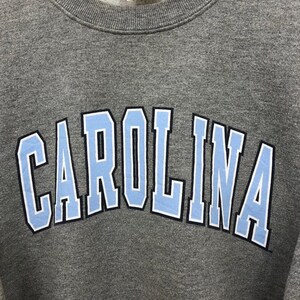 May include: A gray sweatshirt with the word "CAROLINA" in light blue block letters outlined in black and white. The sweatshirt has a crew neck and a tag at the top.