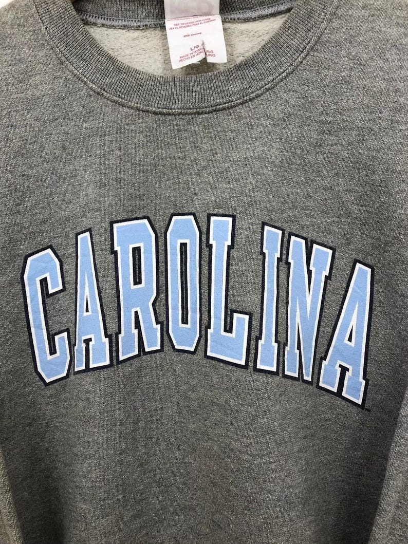 May include: A gray sweatshirt with the word "CAROLINA" in light blue block letters outlined in black and white. The sweatshirt has a crew neck and a tag at the top.
