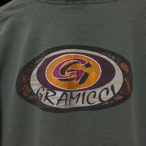 K&ouml;nnte beinhalten: Dunkelgr&uuml;nes T-Shirt mit einem ovalen Logo im Used-Look. Das Logo zeigt das Wort "GRAMICCI" in Wei&szlig; und ein stilisiertes "G" in Lila und Gold. Der Hintergrund ist braun und grau.