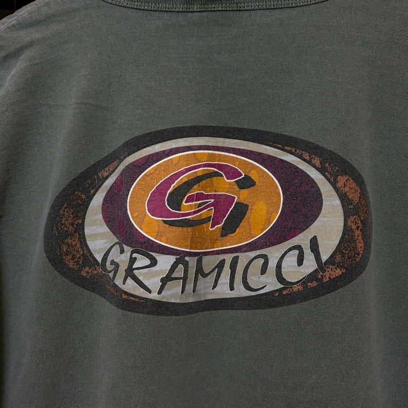 K&ouml;nnte beinhalten: Dunkelgr&uuml;nes T-Shirt mit einem ovalen Logo im Used-Look. Das Logo zeigt das Wort "GRAMICCI" in Wei&szlig; und ein stilisiertes "G" in Lila und Gold. Der Hintergrund ist braun und grau.