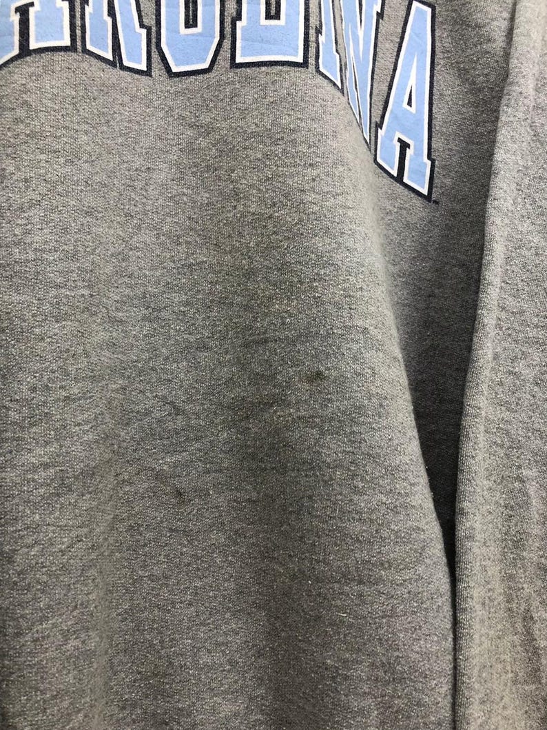 May include: A gray sweatshirt with the word "CAROLINA" in light blue block letters across the top. The letters have a white outline. The fabric appears to be a soft, textured material.